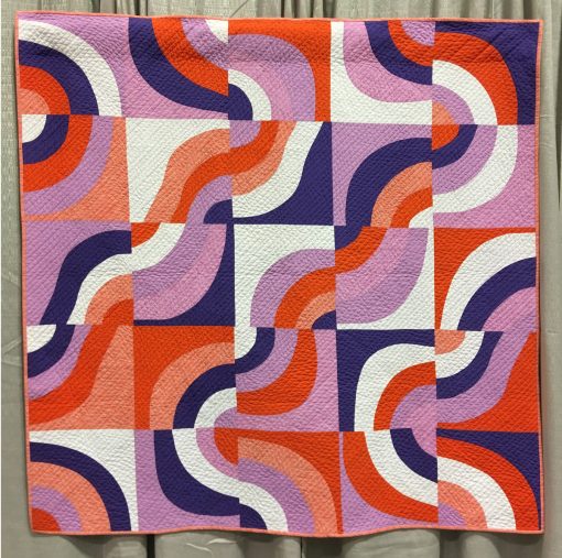 Modern quilt featured at QuiltCon 2019 — “Purple Haze” by Daniela O’Connell @blockMquilts Statement: “This quilt was made taking part in the Pantone Quilt Challenge 2018 using Ultra Violet as a featured colour. I combined white, a bright orange and a light violet to achieve a vibrant colour combination. For the first time I used curved piecing, all curves are cut without the use of a ruler or a template. Individual blocks are turned in different directions to create an interesting overall design. The simple straight line quilting is to support the diagonal direction the design.”