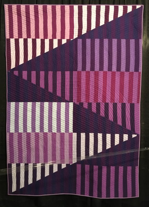 "Ramps" by Elizabeth K. Ray Statement: "I designed Ramps for the 2018 Pantone Ultra Violet Quilt Challenge as part of my series on exploring pieced strips cut into half square triangles and gentle curves. Ramps doesn't use curves. I liked the design element with the sharp extending line and used varying hues of purple and violet along with value contrasts to define strong geometric divisions. My goal was to create clear contrasts and crip points bouncing your eye up and down the ramps. The vertical strips are not intended to mach up by act as pieced areas." [Design Source: Original Design | Techniques: Machine pieced and quilted] displayed in the 2019 Modern Quilt Showcase sponsored by the Modern Quilt Guild at the International Quilt Festival in Houston