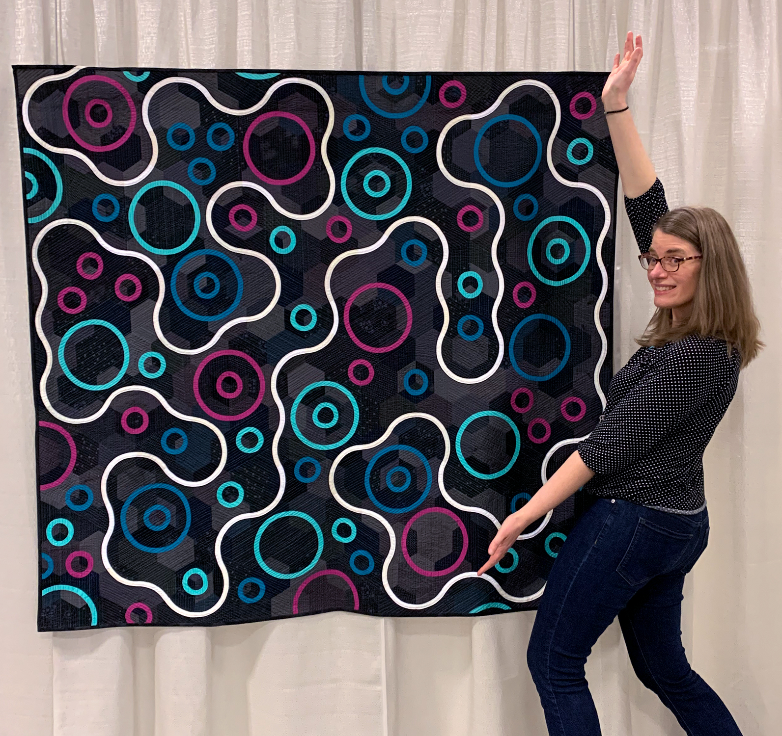 modern minimalist quilt with a dark background and a white curvy line moving around magenta, blue, and aqua circles with the designer and quilter in front of it