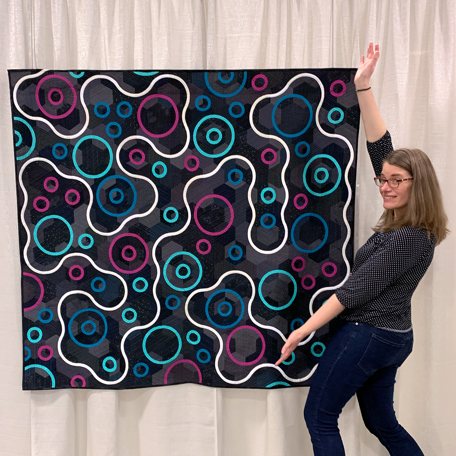 modern minimalist quilt with a dark background and a white curvy line moving around magenta, blue, and aqua circles with the designer and quilter in front of it