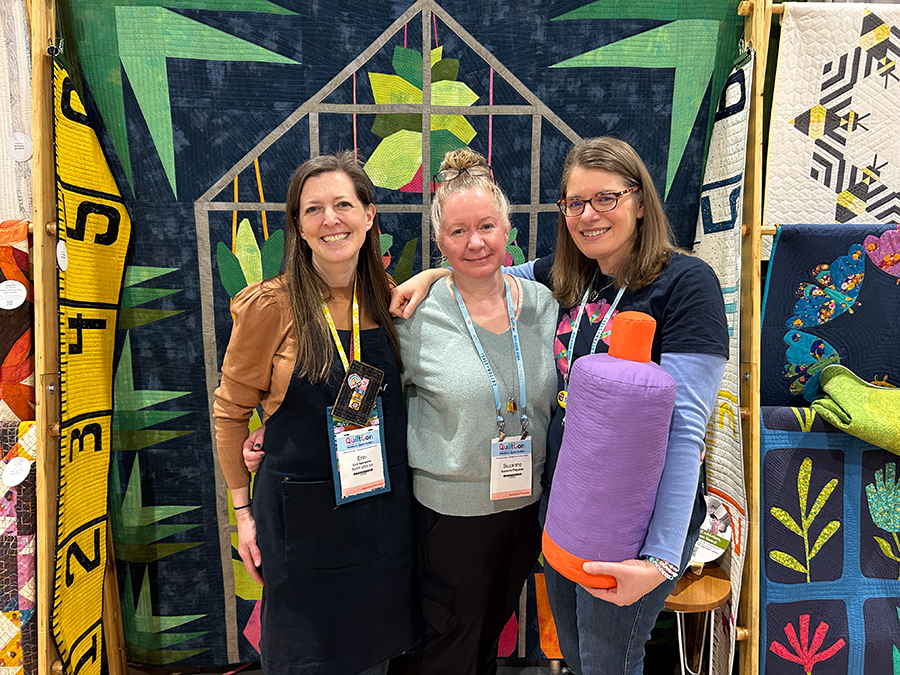 three women smiling and holding a giant spool of thread in front of a quilt