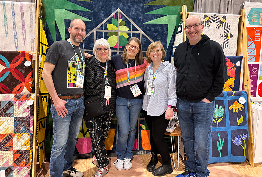 five people standing in front of a quilt smiling