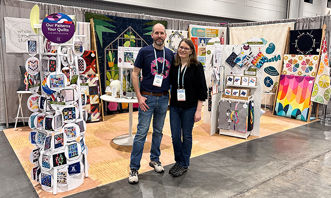 man and woman standing in front of colorful trade show booth with quilts