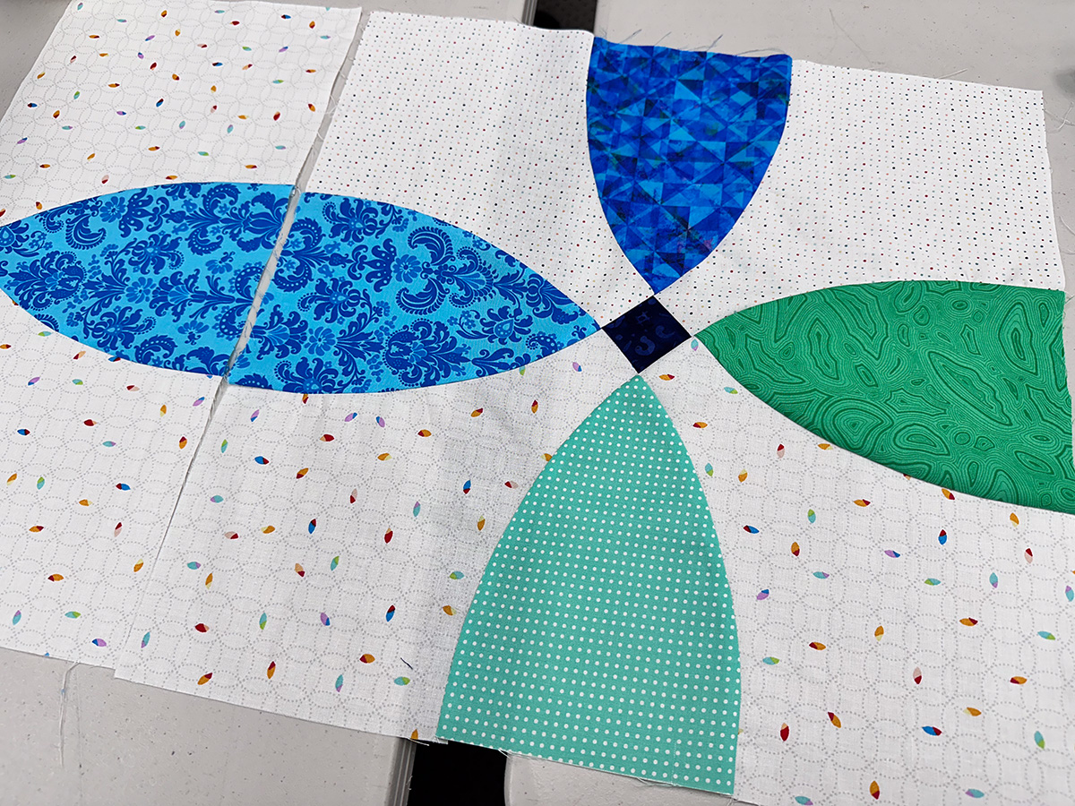 two curved quilt blocks with blue, aqua a green petals and a white background with specks of color