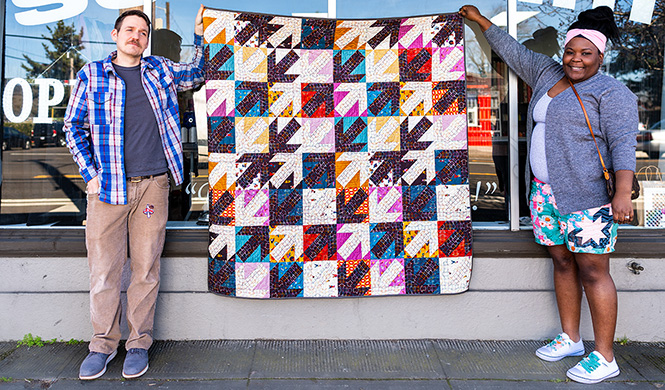 Two people holding up a modern, arrow quilt in front of a storefront