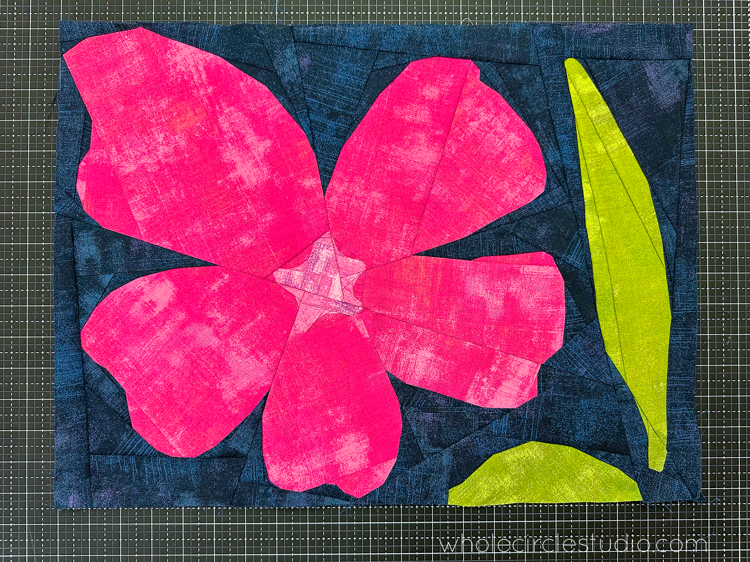 magenta and green hibiscus flower quilt block on a navy background sitting on a cutting mat