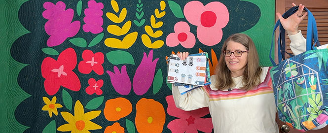 smiling woman holding bags up in front of a colorful flower quilt